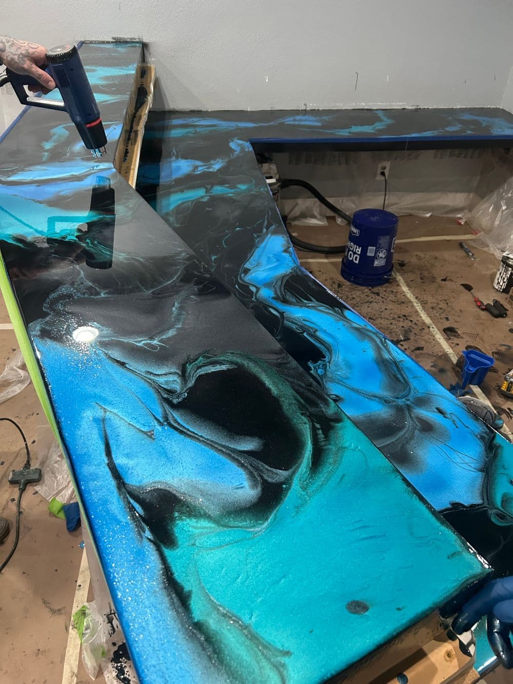 Person applying resin to a vibrant black and blue marbled tabletop in a workshop.